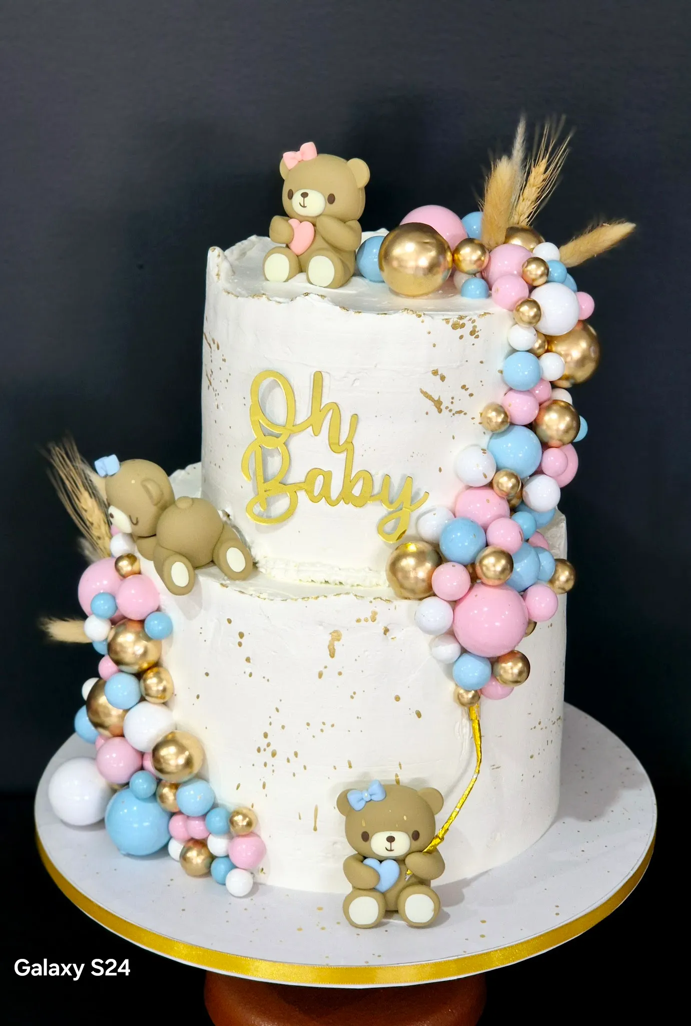 Two-tier white baby shower cake with teddy bears, gold Oh Baby topper, and pastel balloon garland decoration