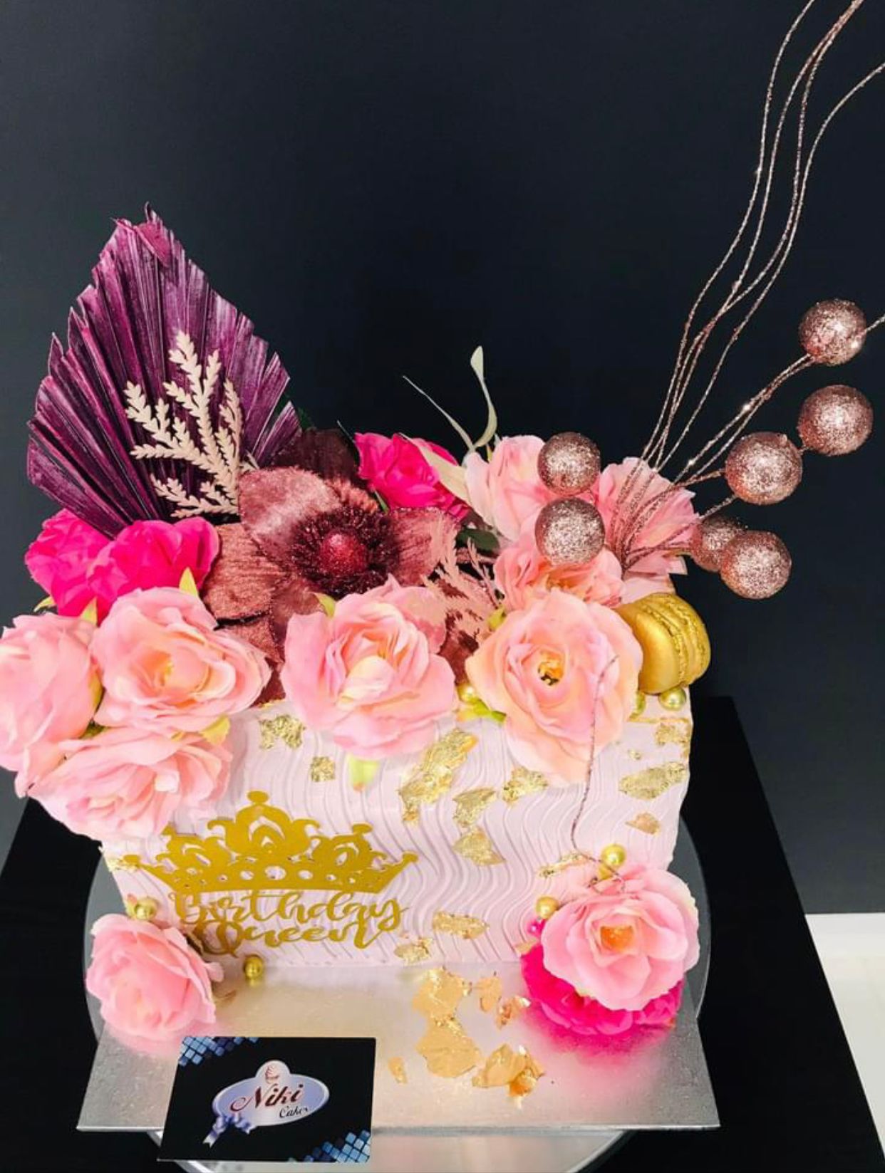 Elegant rectangular birthday cake with gold leaf, dramatic pink and burgundy floral sugar arrangement and crown topper